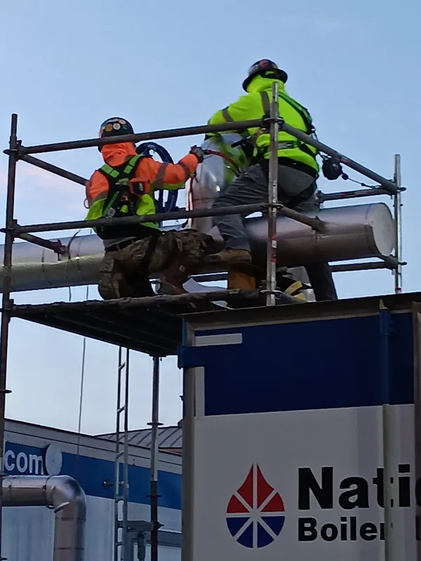Two harnessed insulators jacketing a pipe from a scaffold at dusk